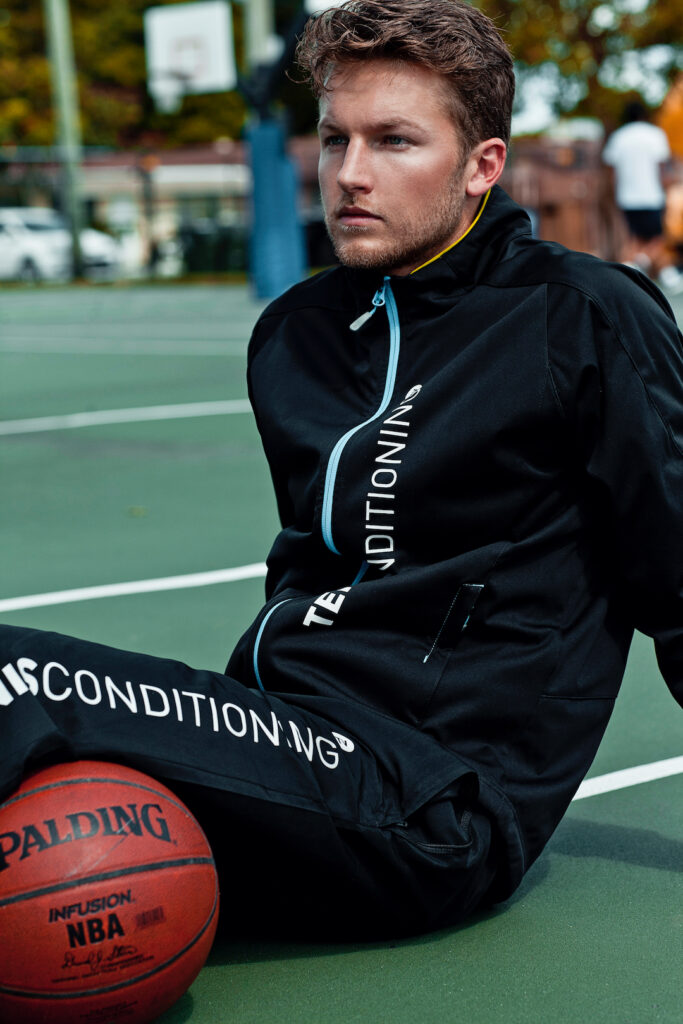 Urban fashion editorial portrait of young man sitting on a basketball court in a dark tracksuit holding a basketball.