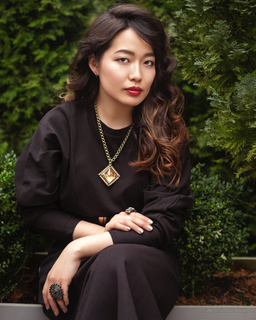 Fashion portrait illustrating how to create editorial fashion portraits with natural lighting with a Japanese woman in a black dress before green hedges.