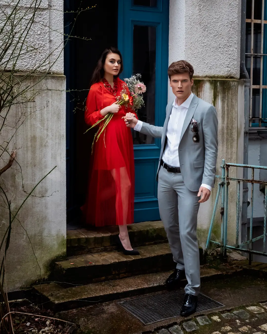Female model in a sheer red dress and male model in a gray suit posing by a blue doorway
