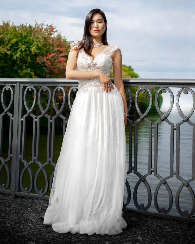 editorial wedding photography. Portrait of elegant Asian woman with long black hair, beautiful makeup standing on a bridge with a water background wearing a white wedding dress and jewelry.