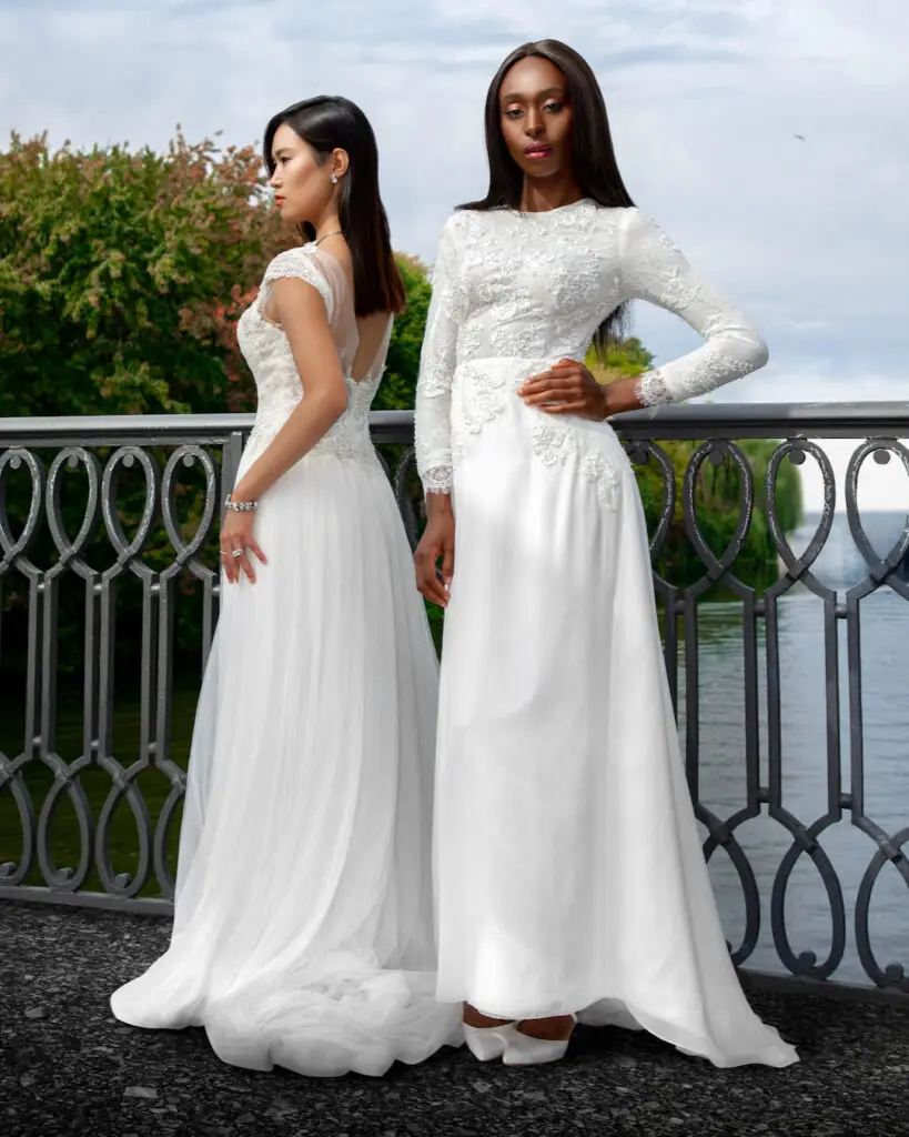 editorial wedding photography. Portrait of two women, one Asian and one Black, both wearing white wedding dresses and high heels, standing gracefully on a bridge with water behind them.