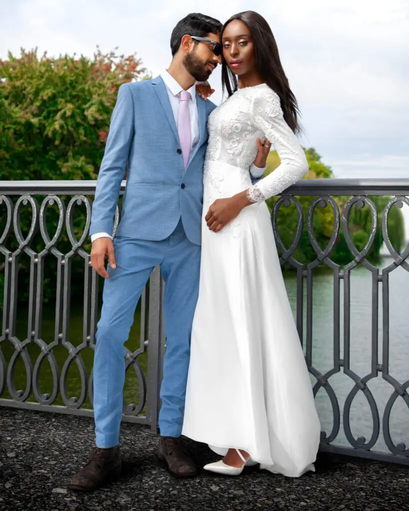 editorial wedding photography. Portrait of an interracial couple — woman in white wedding dress, man in blue suit — standing on a bridge over water.