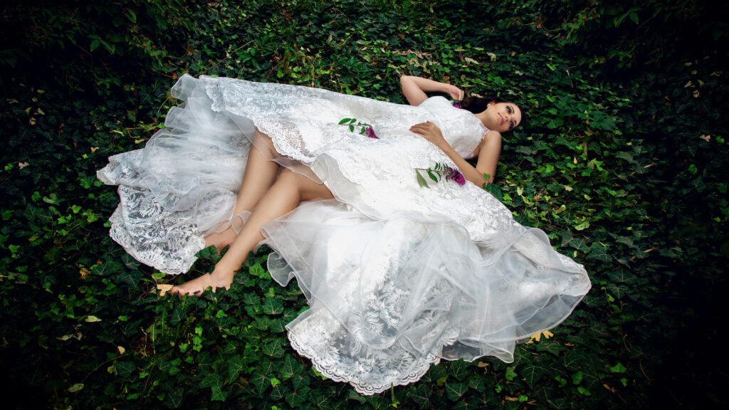 editorial wedding photography. Portrait of a sensual white woman with long natural curly dark brown hair, beautiful makeup, lying outdoors on a bed of green leaves wearing a white wedding dress.