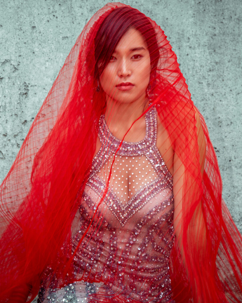 editorial wedding photography. Close-up of an Asian woman with a red veil and evening dress in front of a concrete wall — an ode to creative wedding portraits.