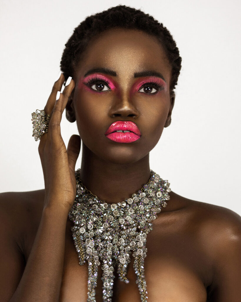 Beauty editorial portrait of young Black woman with short black hair, pink lipstick, manicured nails, and diamond necklace against bright background.