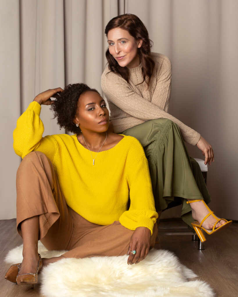 Clothing brand photoshoot lifestyle portrait: sensual Black woman lounging on floor with white woman on bench wearing casual clothing and heels
