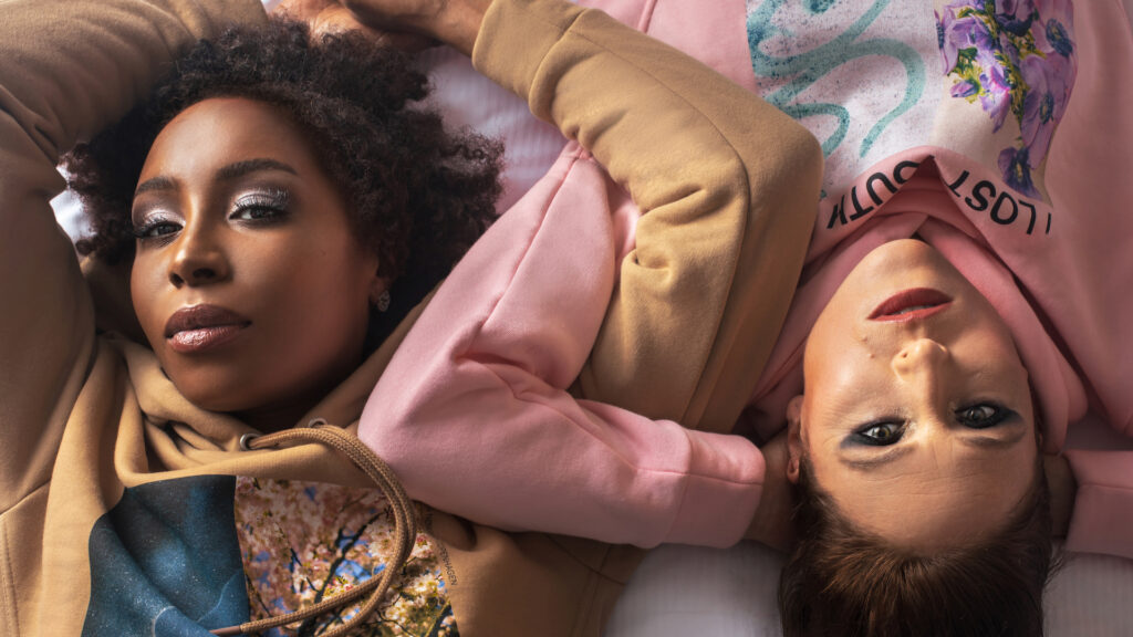 Clothing brand photoshoot closeup: sensual Black woman face-up on bed intertwined with white woman wearing colorful hooded pullovers