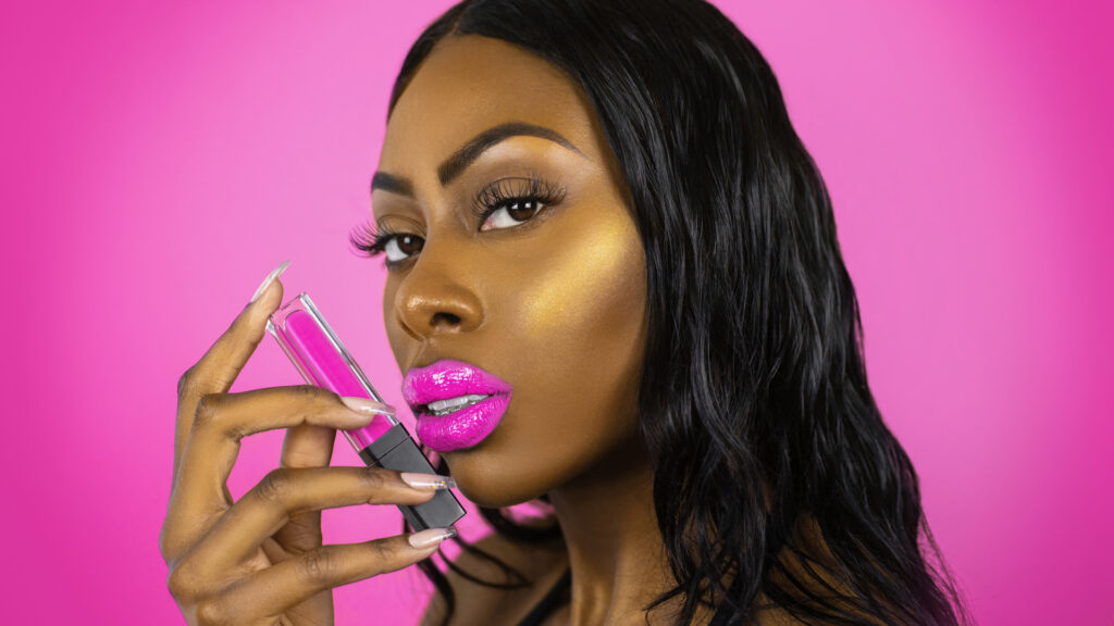 Artistic beauty photography portrait of a young Black female with long black hair holding a pink lipstick in front of a pink background.