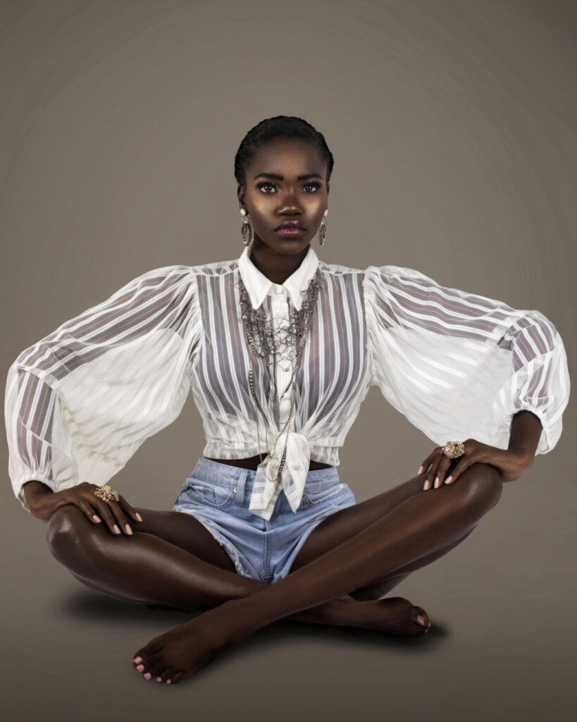 A studio portrait of a serene young black female sitting on the floor with short black hair and pink nails wearing a white sheer blouse with denim shorts and silver jewelry.