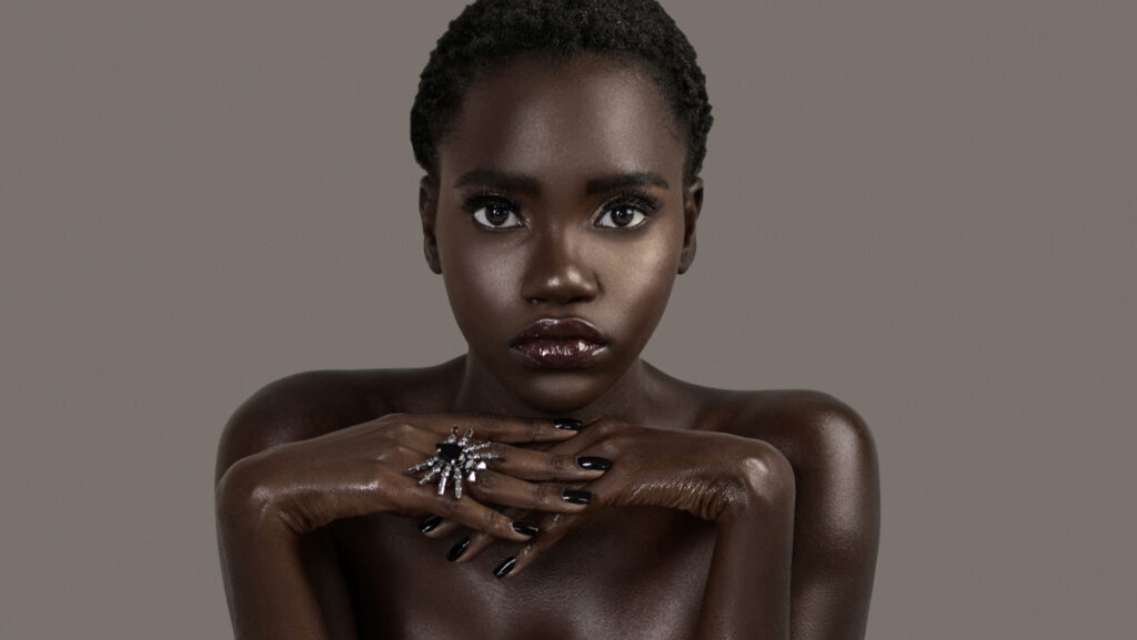 A portrait of a serene young black female with short black hair, moist lips, perfectly black manicured nails with silver jewelry looking straight at the camera.