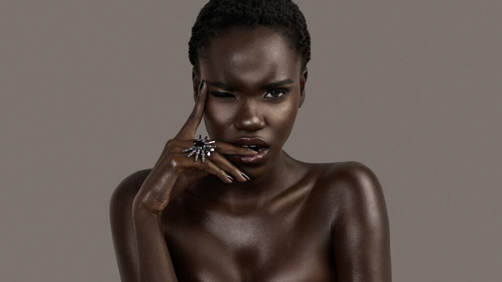 A portrait of a serene young black female with short black hair, moist lips, perfectly black manicured nails with silver jewelry looking straight at the camera.
