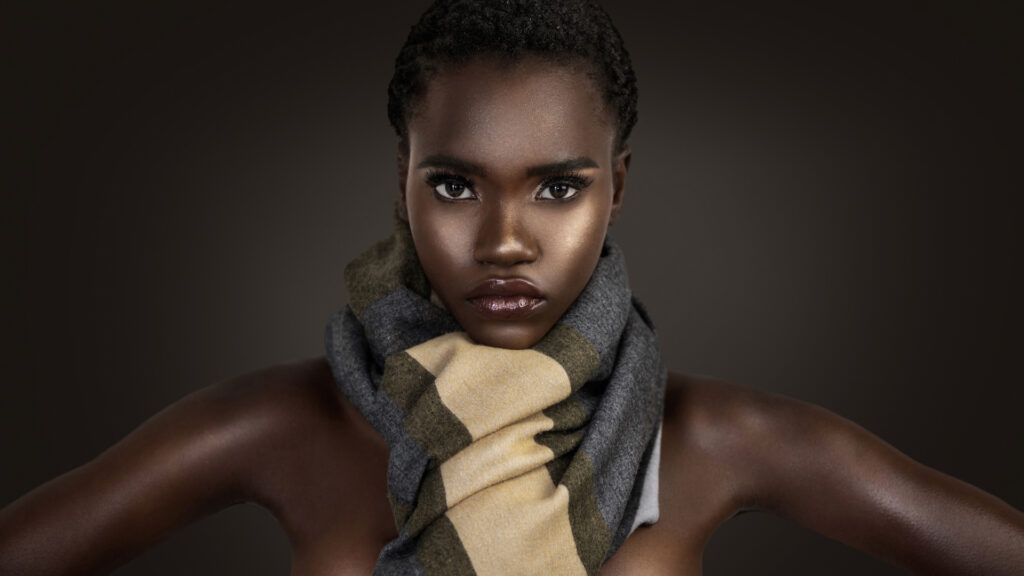 Beauty editorial portrait of young Black woman wearing colorful scarf against dark background.