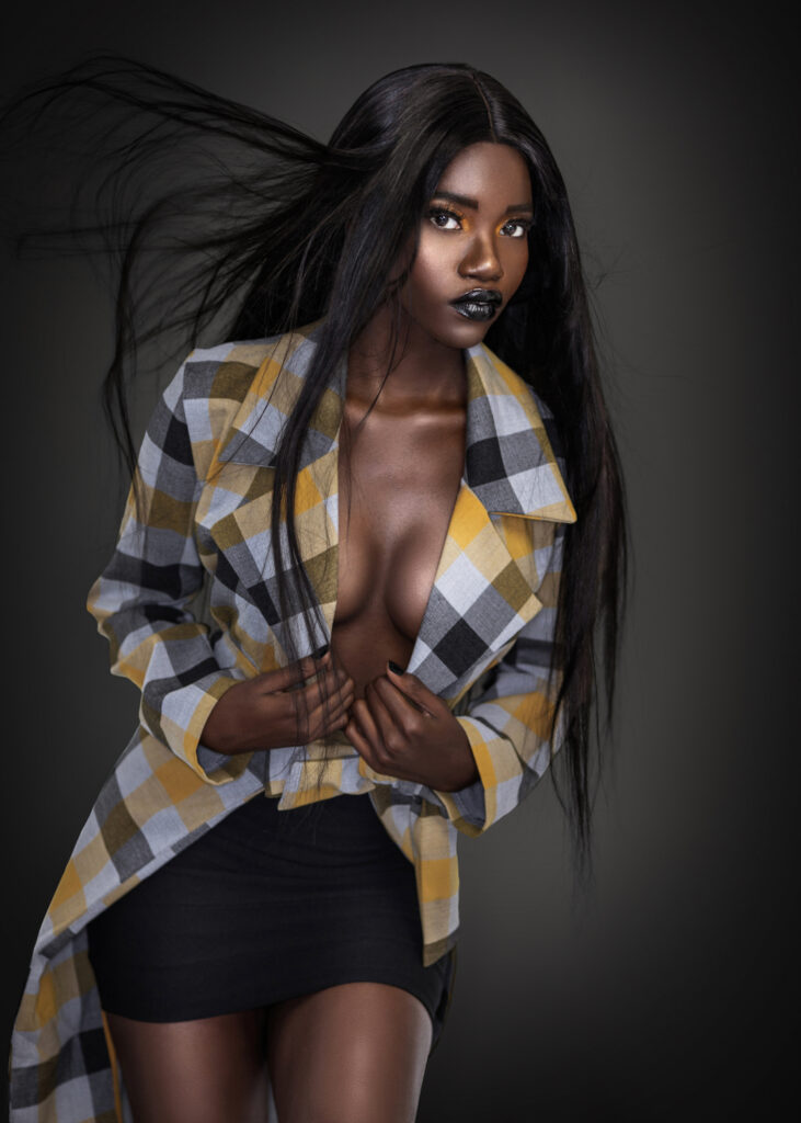 Serene beauty editorial portrait of young Black woman with long hair, dark lipstick, colorful coat, and black skirt against dark background.