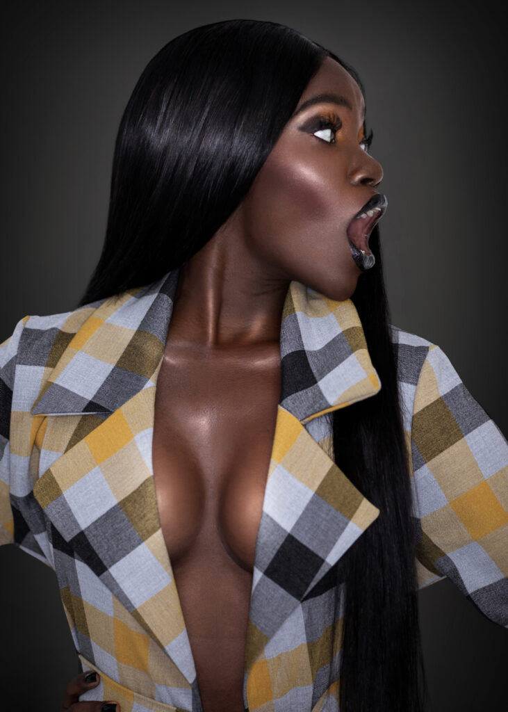 Serene beauty editorial portrait of young Black woman with long hair, dark lipstick, colorful coat, and black skirt against dark background.