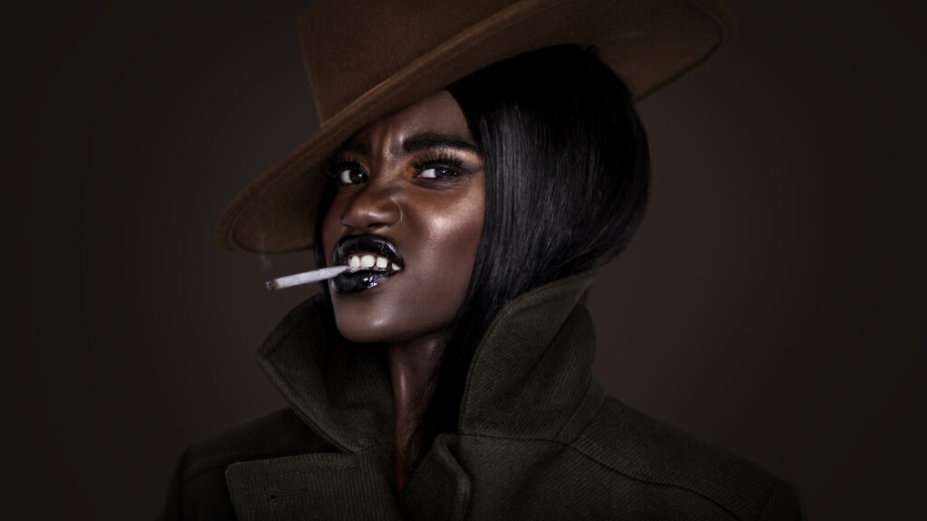 Portrait of a smoking young black female with long black hair, beautiful makeup, popping dark lip stick posing by herself wearing a dark green coat & brown hat in front of a dark background.