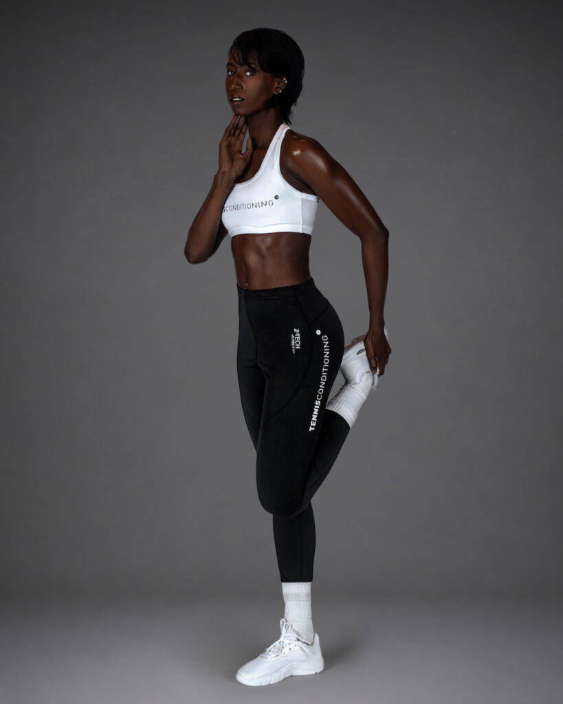 Gigi performing a standing quadriceps stretch in a white sports bra and black leggings