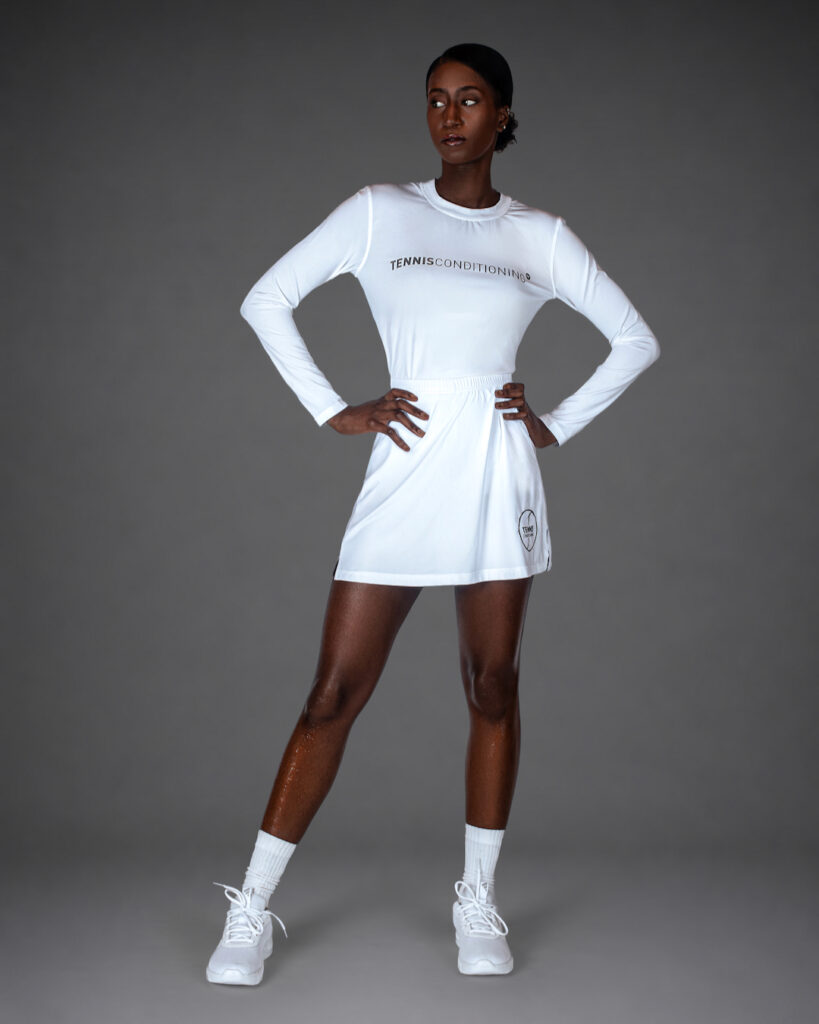 Gigi standing in an all-white tennis-inspired athletic outfit against a gray studio background