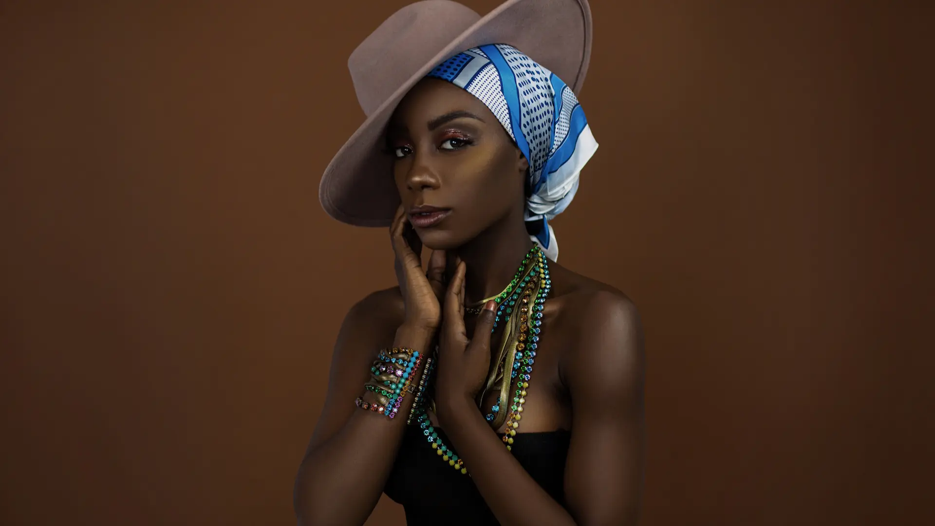 Black model wearing a blue-and-white headscarf, beige hat and colorful jewelry