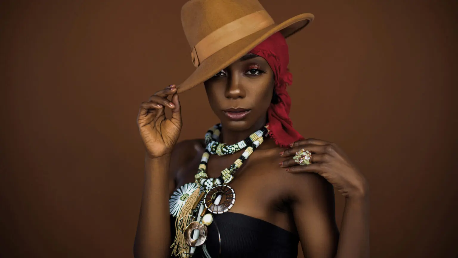 Black model wearing a red headscarf, brown hat and colorful necklace