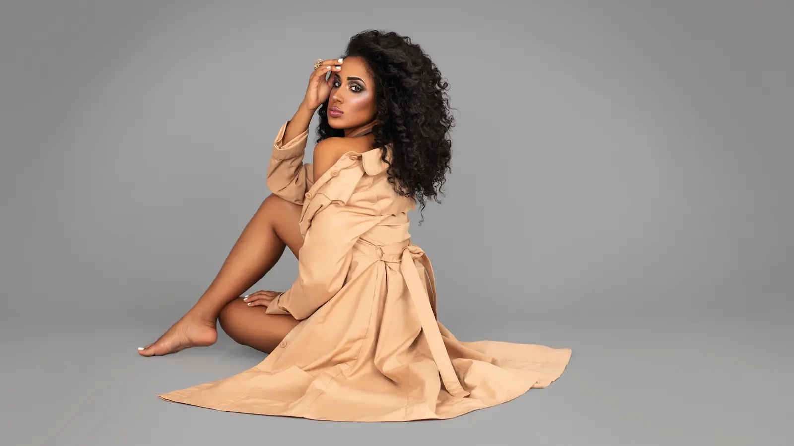Curly-haired model in a beige trench coat seated on a grey studio floor