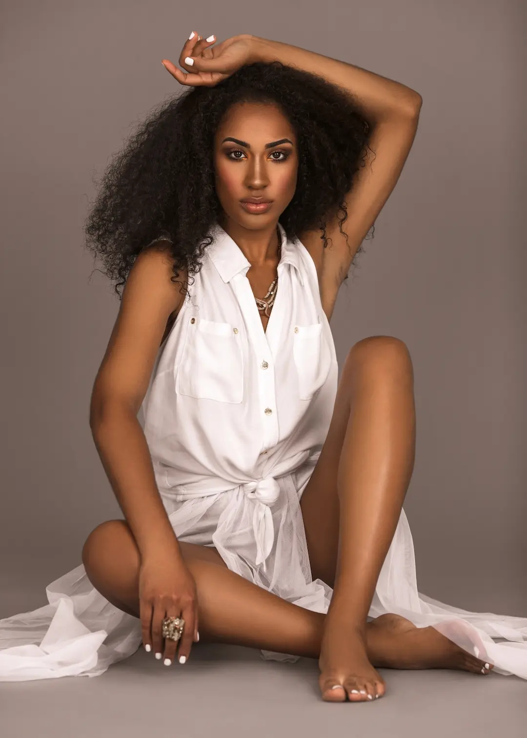 Willina Rodriguez seated barefoot in a flowing white dress for the Curls Poppin curly hair beauty editorial