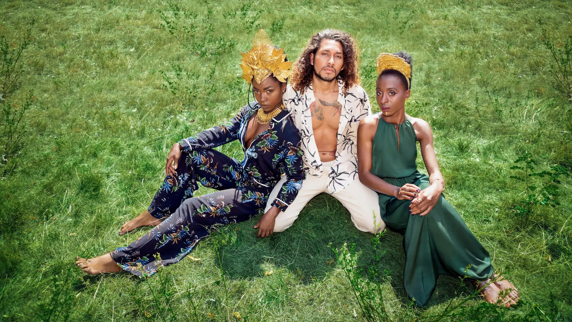 Three models seated together in elaborate African-inspired fashion in a green landscape