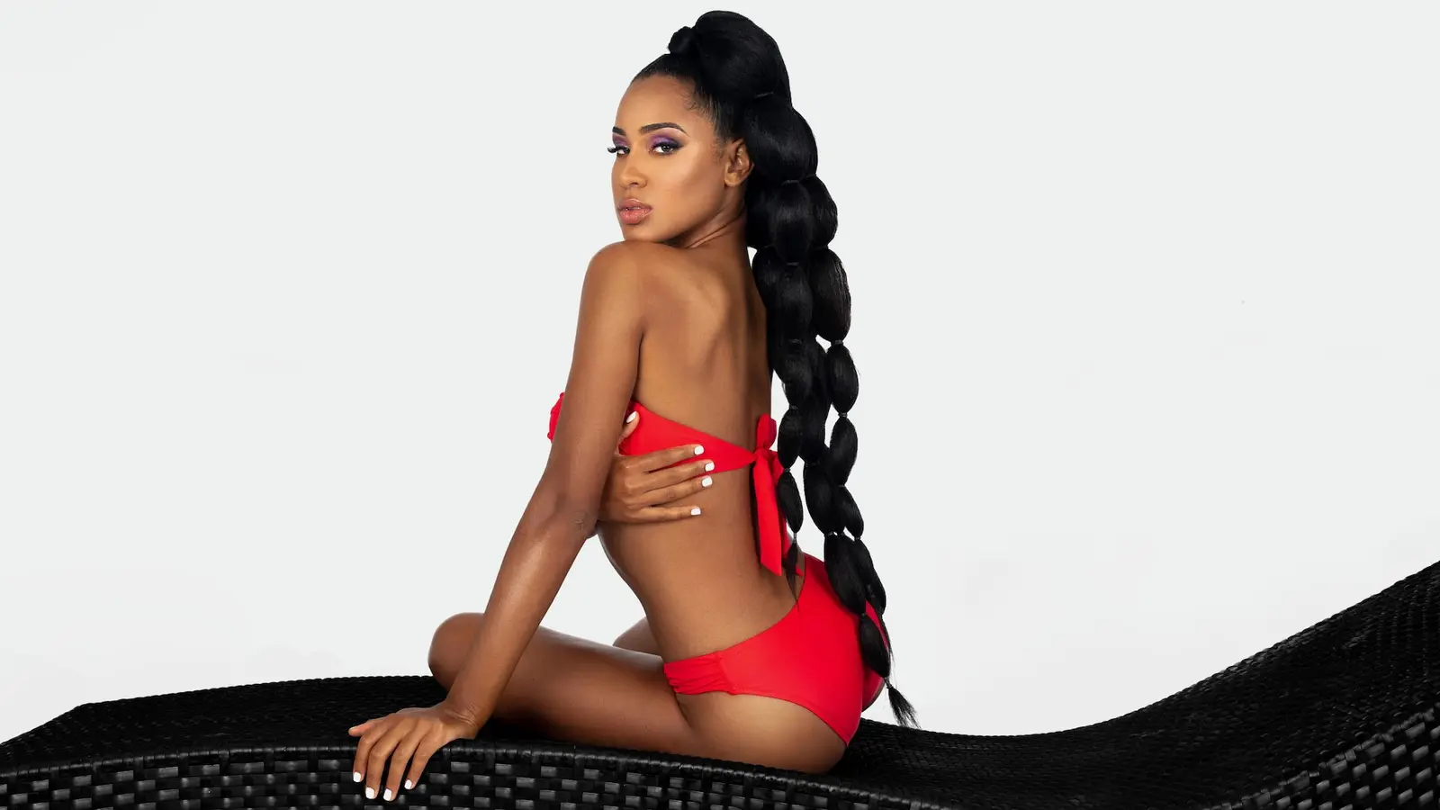 Willina in red swimwear seated on a dark woven bench in a white fashion studio