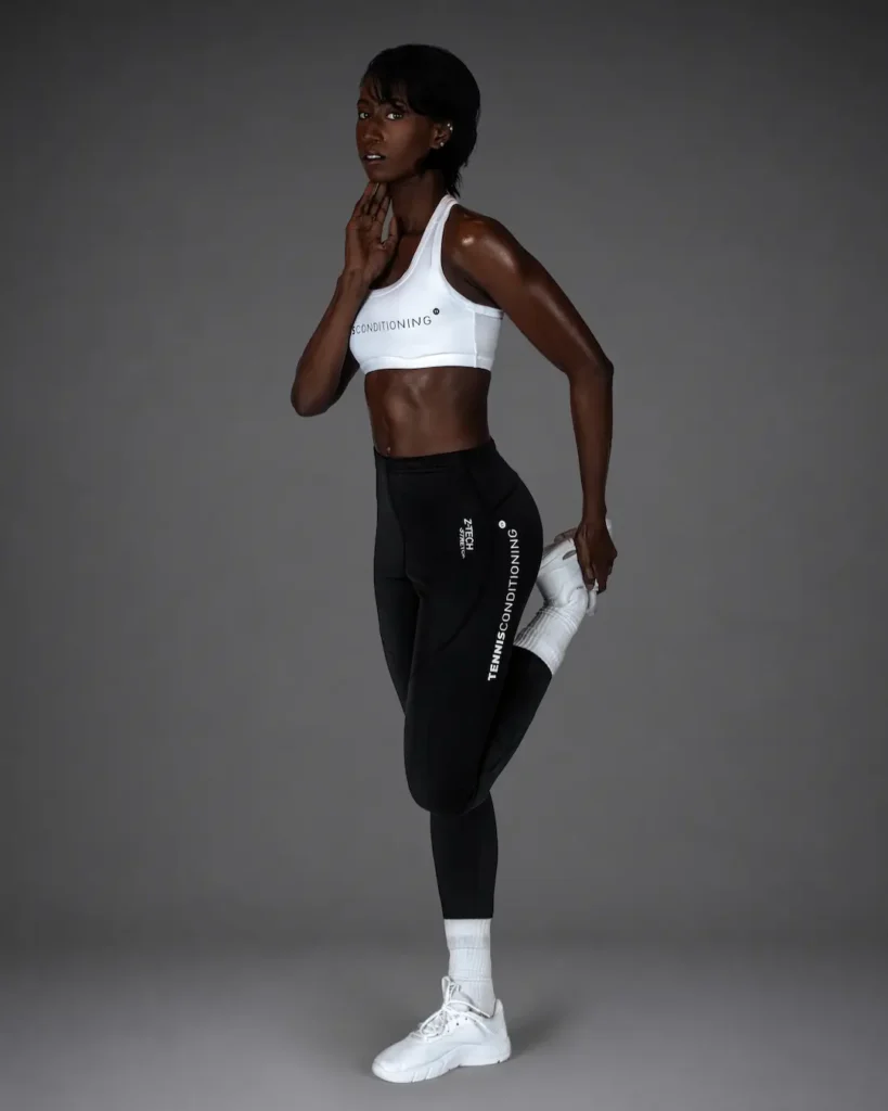 Gigi performing a standing quadriceps stretch in a white sports bra and black leggings