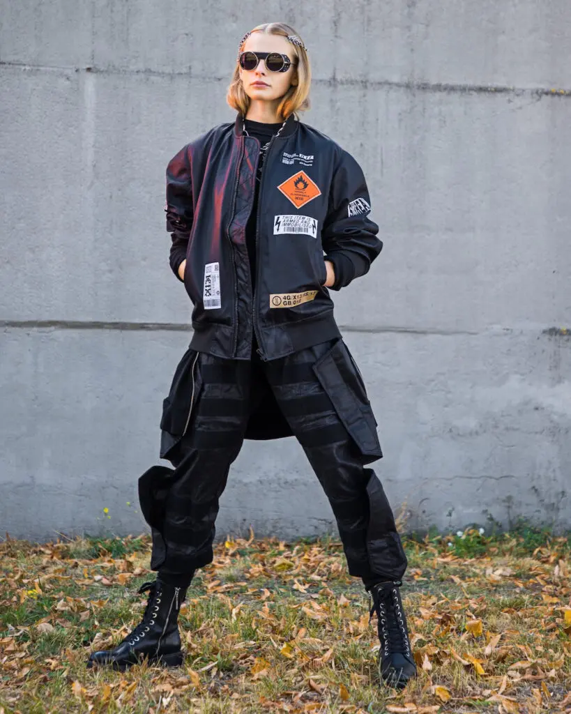White woman in a black jacket, trousers, combat boots and sunglasses in a park
