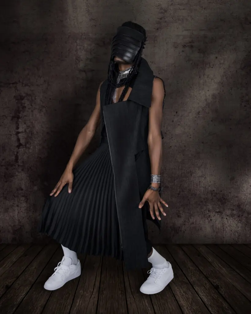Black male model wearing a sleeveless coat, black dress and leather mask