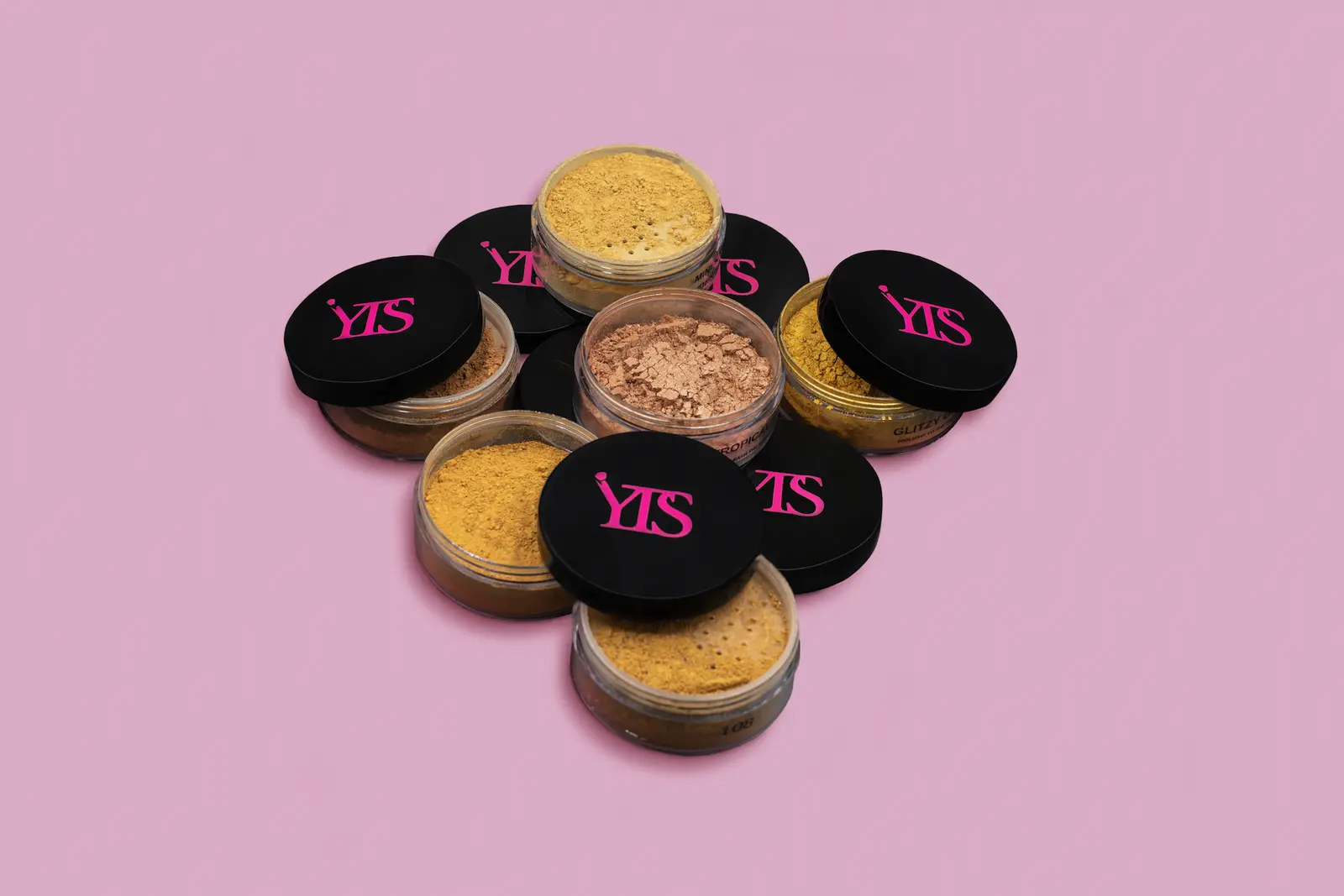 Gold beauty powder jars arranged around lipsticks on a soft pink background