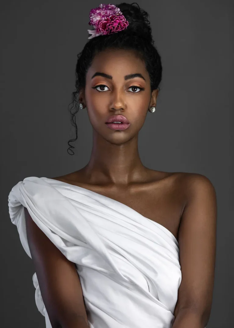 Black model draped in white fabric with flowers in her curls and purple lipstick