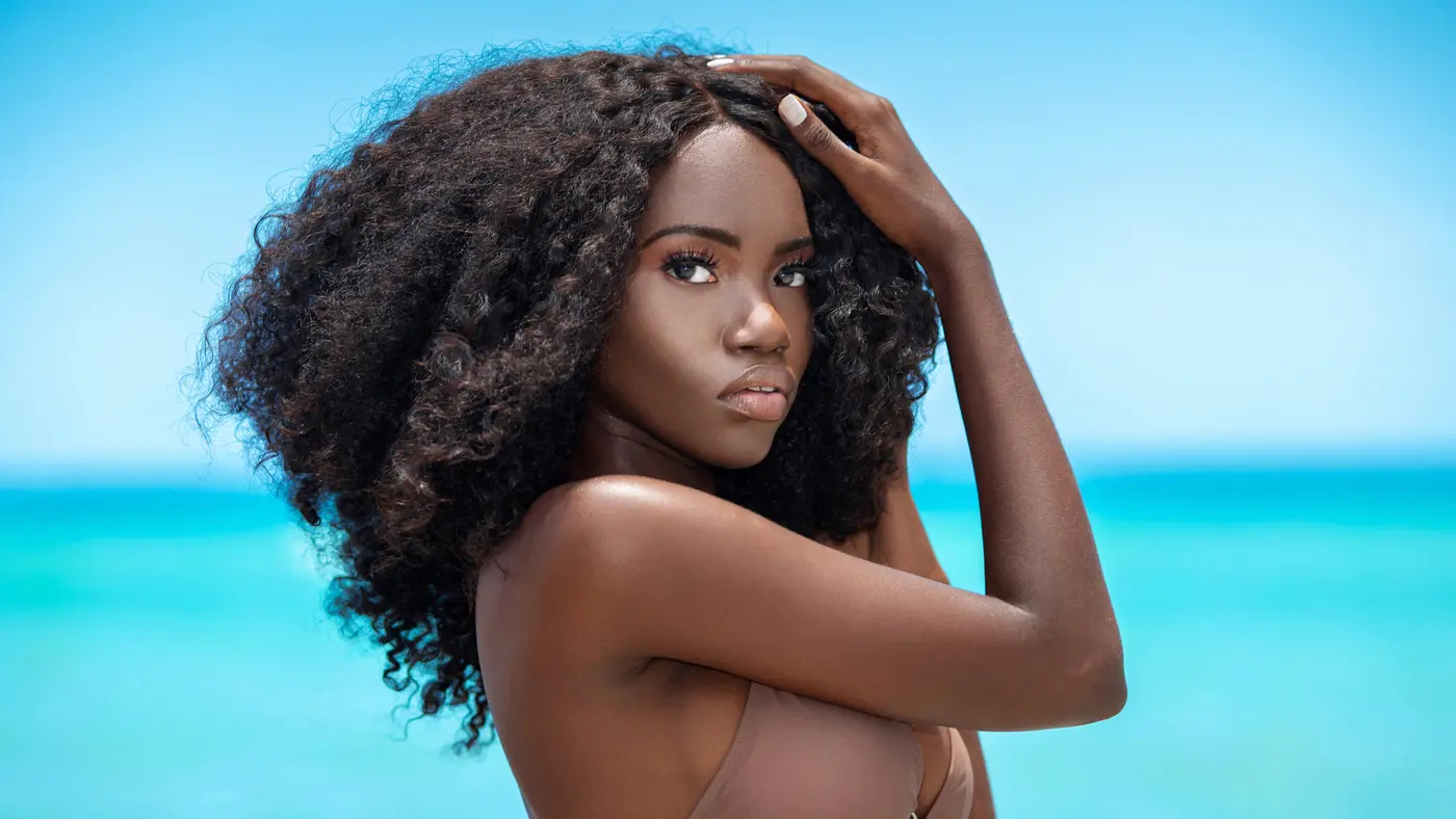 Black model with long curly hair posing in brown swimwear beside the ocean