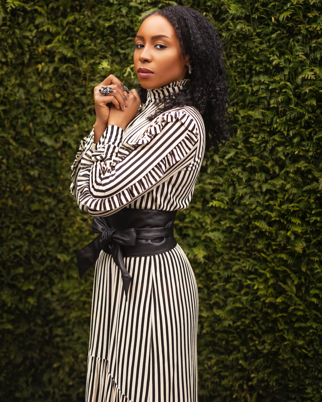 Woman in a black-and-white striped dress photographed beside green hedges