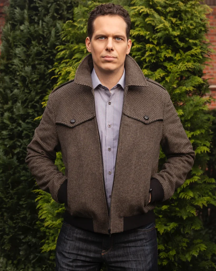 Man wearing a brown jacket and gray shirt in front of green hedges