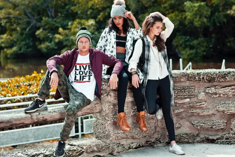 Three models in layered streetwear pose on a stone wall beside a Miami lake.
