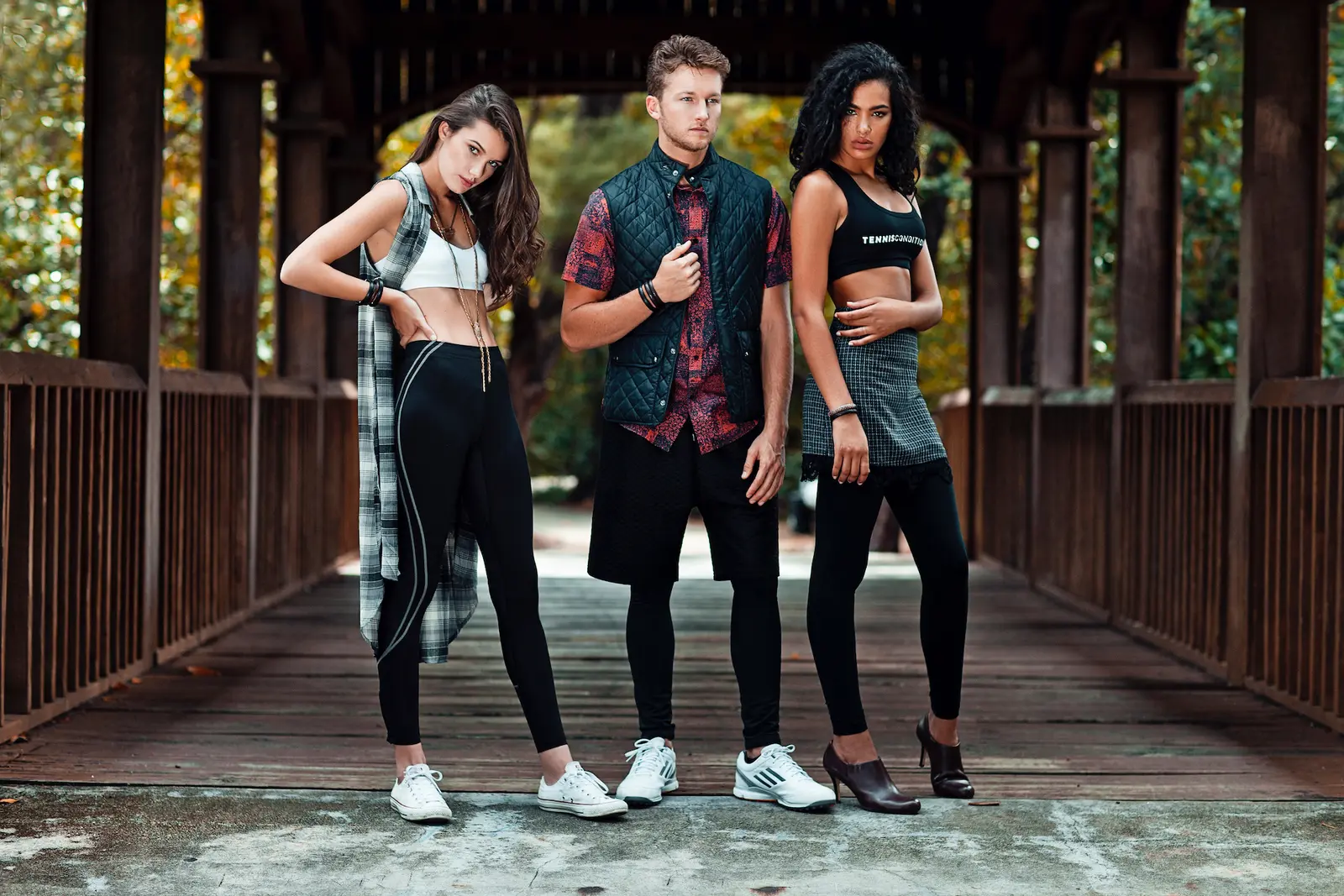 Three models in modern streetwear stand on a wooden bridge in a Miami park.