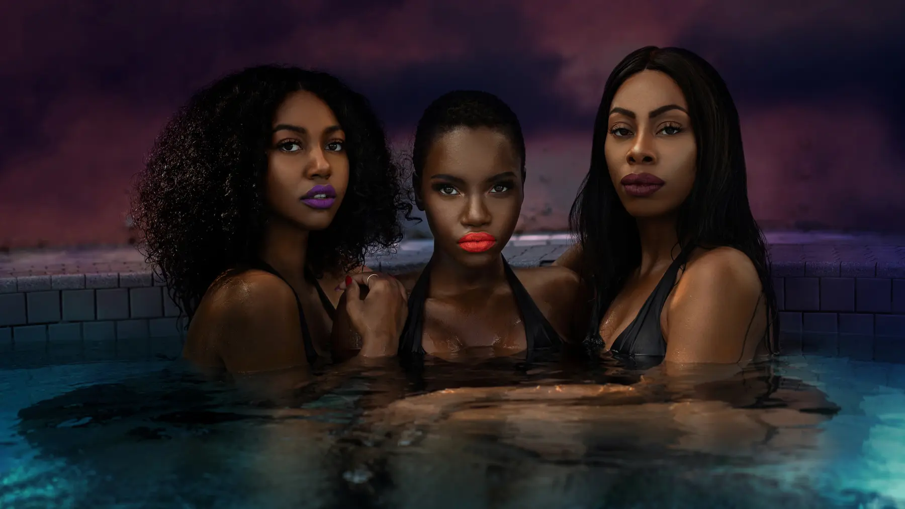 Three Black women wearing bold lipstick and posing together in a swimming pool at dusk.