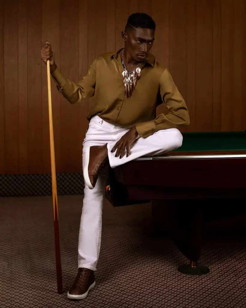 Black male model wearing a brown shirt, white jeans and sneakers in a pool hall