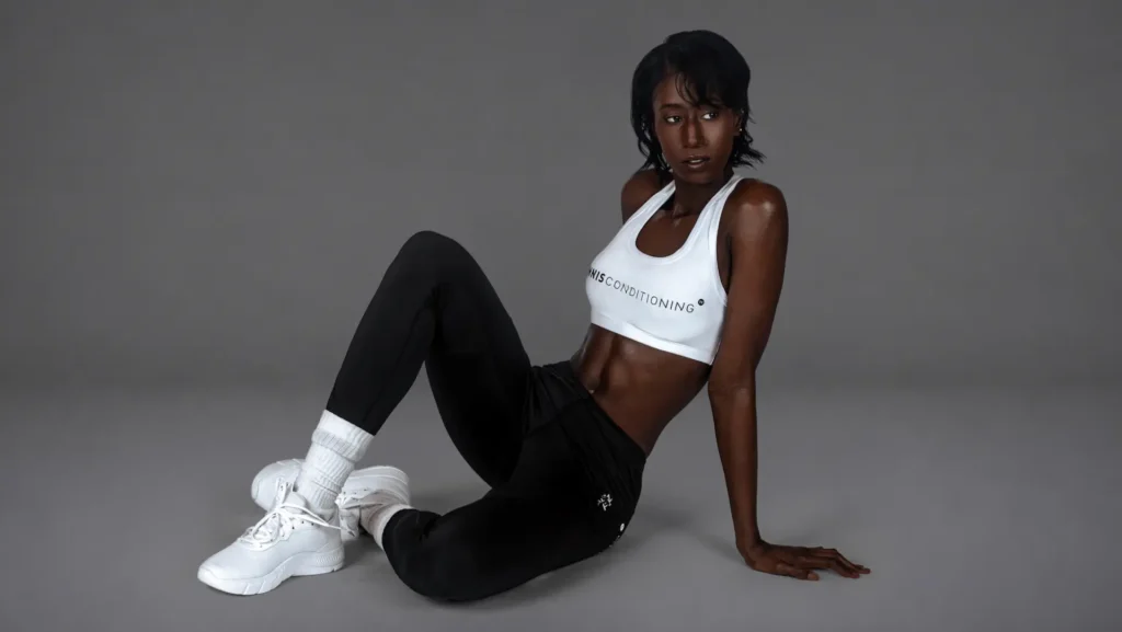 Gigi seated on the studio floor in a white sports bra and black leggings for the Strength in Motion editorial
