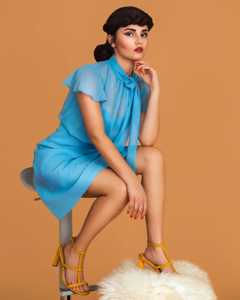 Mariana seated in a bright blue chiffon dress with yellow heels against the brown studio backdrop