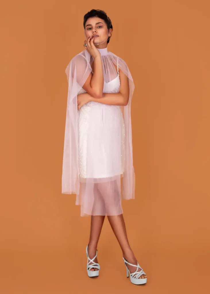 Mariana standing in a layered white dress and silver heels against a warm brown studio backdrop