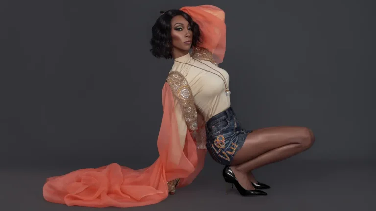 Portrait of Black woman with wavy black hair and makeup standing in a solo portrait inside a studio with grey background wearing short denim skirt with orange dress and black high heels.
