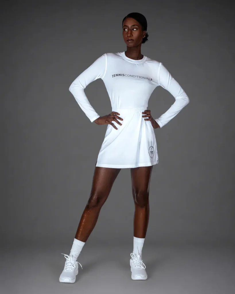 Gigi standing in an all-white tennis-inspired athletic outfit against a gray studio background